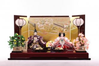 溜塗の屏風は京金彩加工を施した御所庭園鞠桜の絵柄を上品に、飾台は、高級感のある高さと二色塗分けが施され、お雛様がより豪華に演出されています。
花は京都御所にある左近の桜、右近の橘をまねた物と言われています。