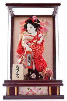 赤い絞りと金彩花柄を着物の半身ずつにあしらっています。花梨色の木製枠のアクリルパノラマケースなので、3方向から羽子板を遮らず美しく魅せます。背面とケース下部の幕板には桜模様の刺繍を施しました。家庭円満の象徴である大きな御殿鞠が彩りを添えています。